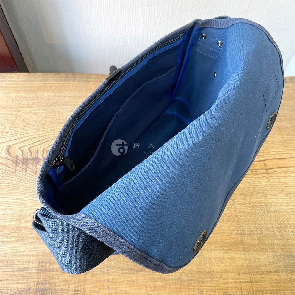 藍色純棉帆布雙扣郵差包，特寫包內結構，有多個隔層，可見拉鍊和金屬扣。