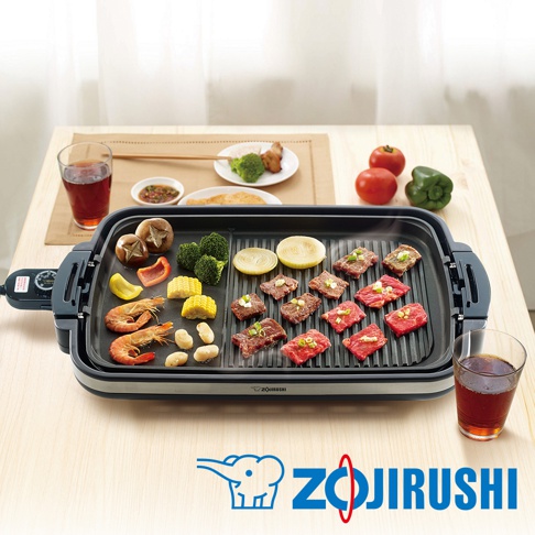 分離式電烤盤上烤著牛肉片、鮮蝦、玉米、香菇、青椒、洋蔥和花椰菜，烤盤為黑色，搭配銀色邊框和黑色把手，左側有黑色控制面板。