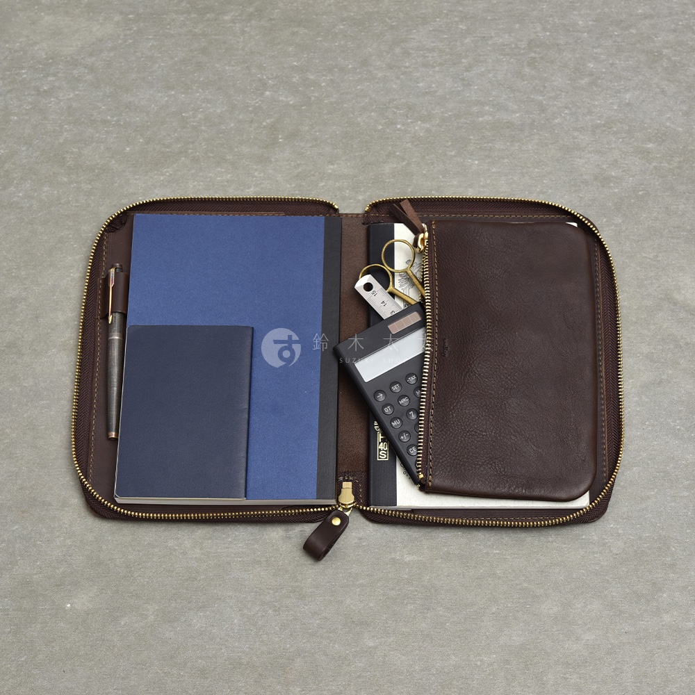 深棕色皮革收納包，打開後左側有藍色筆記本、深藍色小冊子和一支筆，右側有計算機、剪刀和另一本筆記本，拉鍊為金色。