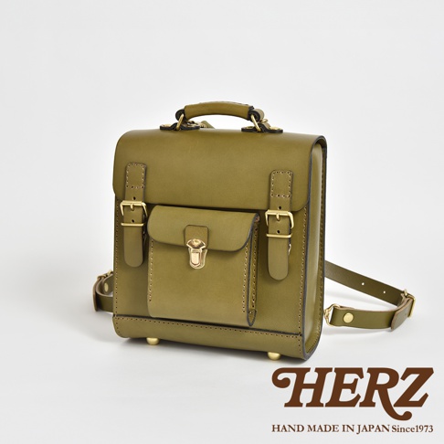 橄欖綠皮革學院風迷你後背包，正面有翻蓋口袋及金屬扣鎖，兩側有帶金屬扣的皮帶裝飾，頂部有提把，底部有金屬腳釘，整體為簡約經典設計。