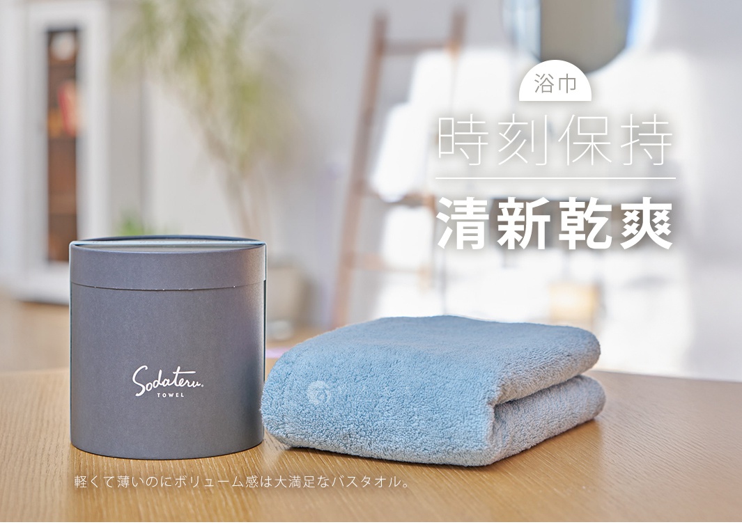 灰色圓筒形包裝盒旁，擺放著一條淺藍色、對折的柔軟毛巾，毛巾表面有細緻的絨毛紋理。