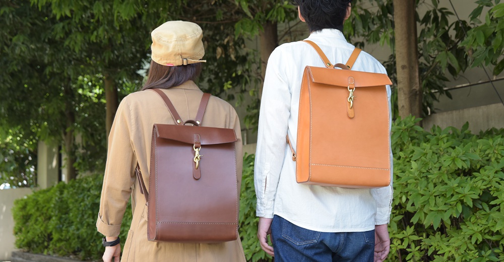 兩款職人手作皮革後背包，一款為深棕色，另一款為淺棕色，皆為方形翻蓋設計，配有金色金屬扣環與皮革提帶。