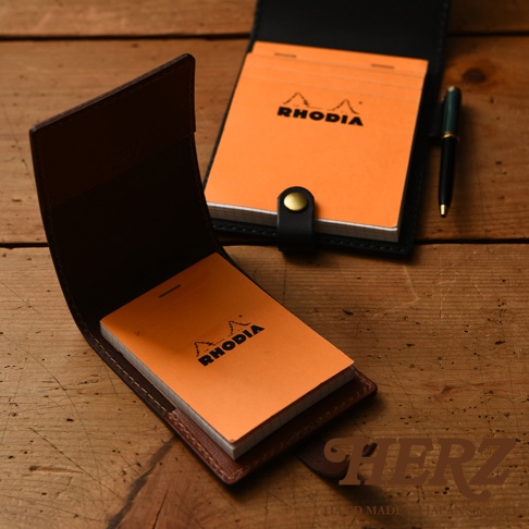 深棕色皮革製成的職人手作便利貼收納套，內含橘色 RHODIA 筆記本，另一黑色皮革收納套內也放有橘色 RHODIA 筆記本，其中一本筆記本旁有一支綠色筆。