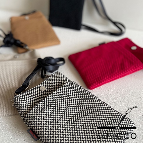 三河木綿刺子織手機包，黑白格紋款式，搭配黑色織帶與釦子。背景為淺米色、卡其色、紅色與黑色手機包。
