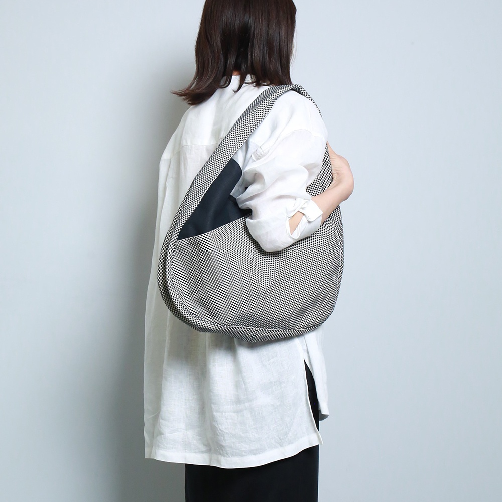 一名女性身穿白色長袖上衣，斜挎一個半月形刺子織斜背包。包包以黑白細小格紋圖案為主，搭配一塊黑色拼接，肩帶與包身同為黑白格紋。