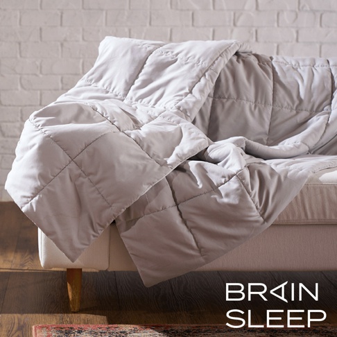 一張淺灰色雙人調溫被，表面有菱形格紋縫製，被子鋪蓋在米白色沙發上，背景為白色磚牆。