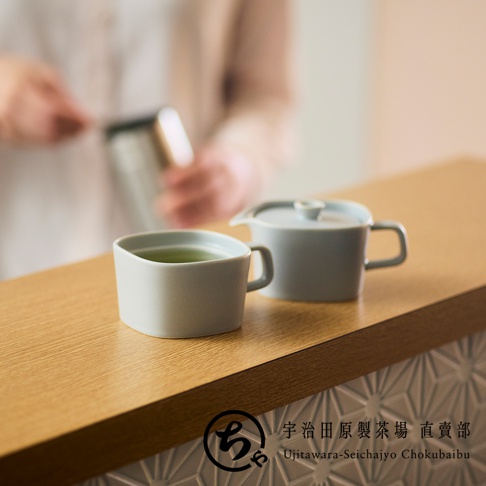 在淺色木質吧台上，擺放著一組宇治田原製茶場的淡雅灰色美濃燒茶具，包含一個裝有翠綠茶湯的方形馬克杯以及一個帶蓋的茶壺，背景模糊處隱約可見正在沖茶的人影，右下角印有該品牌的中英文名稱。
