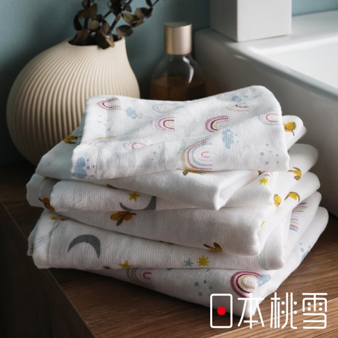 一疊印有彩虹、月亮和星星圖案的白色紗布方巾，置於木質桌面，背景有白色陶瓷花瓶和一瓶化妝水。