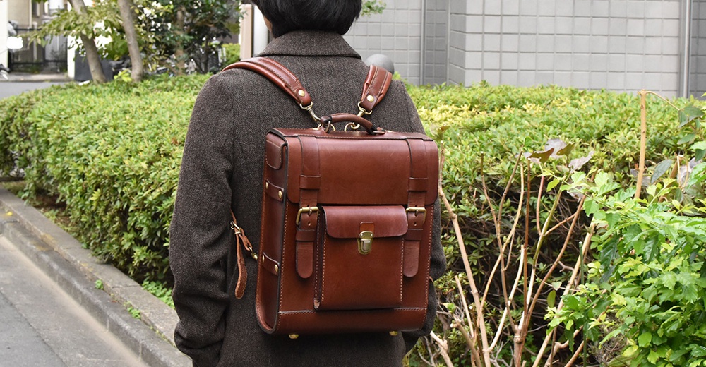 棕色皮革後背包，學院風格設計，有翻蓋、金屬扣環和雙肩帶，適合日常或旅行使用。