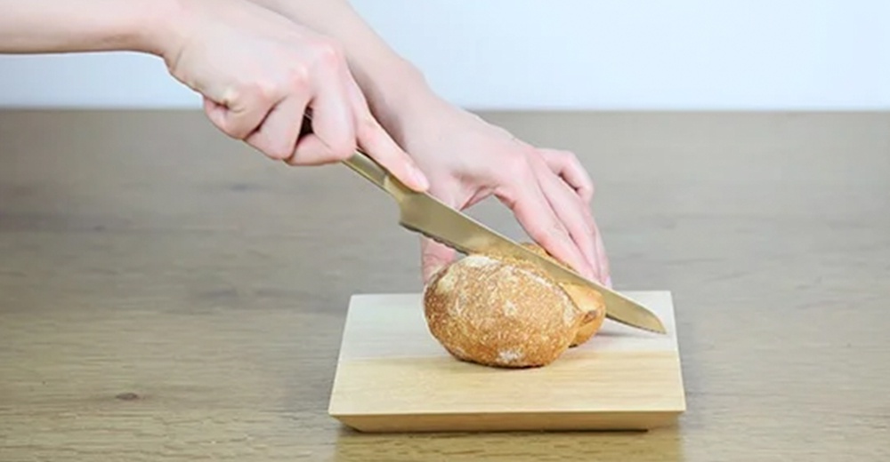 雙手拿著金色麵包刀，正在切著放在淺色木製砧板上的麵包。
