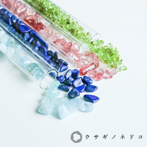 四支透明試管瓶，內含淺藍、深藍、粉紅、淺綠色等不同尺寸、形狀的礦石碎料，散落於瓶外，營造自然質感。
