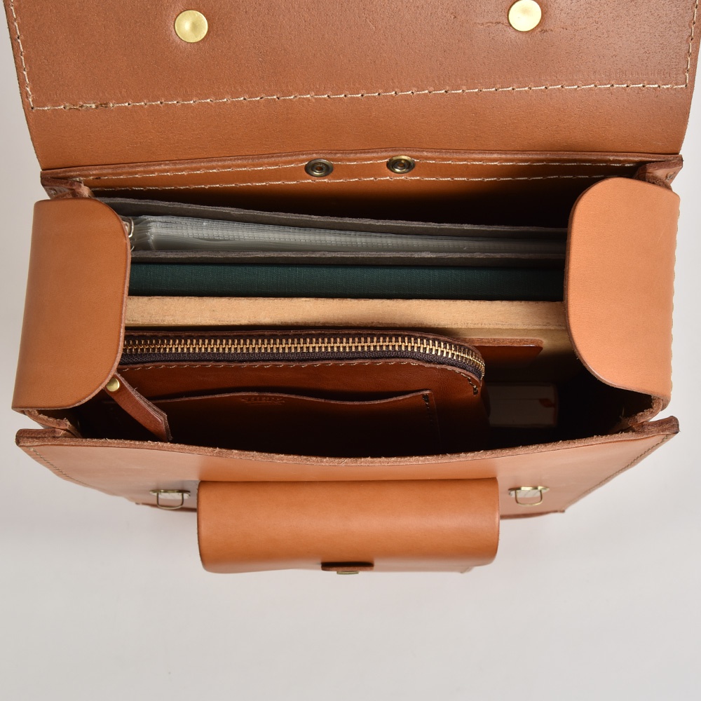 職人手作皮革經典學院風後背包 R-100，展示駝色皮革材質，內部可見文件夾、書本及拉鍊收納袋，設計簡約實用。
