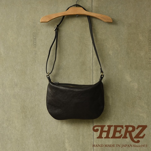 黑色皮革單肩半月包，附有可調式背帶和金色拉鍊，懸掛在木製衣架上。