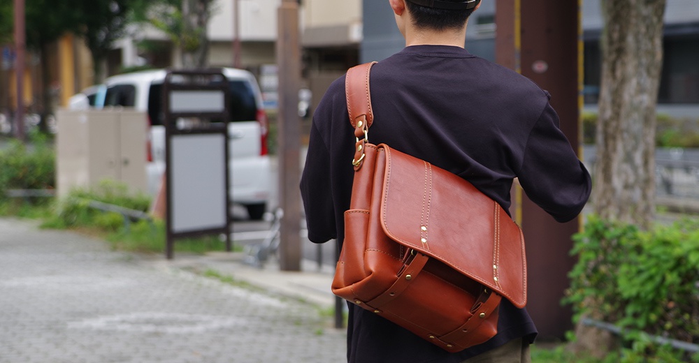 棕色皮革復古郵差包，帶有金屬釦件和縫線細節，由一人斜背於肩上。