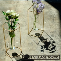 兩個黃銅金屬框架花器，各自裝有玻璃試管，分別插著白色玫瑰和紫色花朵，置於水泥背景上，光影投射。