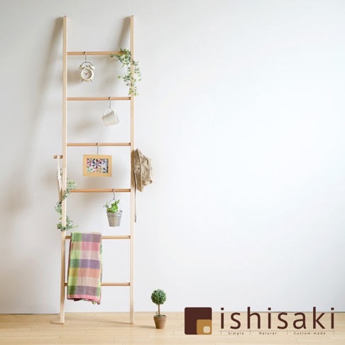 淺木色木質牆面收納架，梯形結構，掛有綠植、鬧鐘、馬克杯、裝飾畫框、帽子和毛毯，架子旁有一盆小型盆栽。