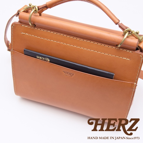 一棕褐色皮革手提側背包，包身設有外插袋，可放入筆記本，並壓印有HERZ品牌標誌及產地資訊。