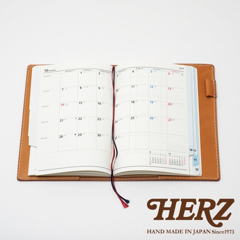 職人手作皮革B6記事本書套，展現天然皮革的溫潤質地，焦糖棕色皮革外殼，內含一本翻開的月曆筆記本，書本裝有紅藍色書籤帶，簡約實用。
