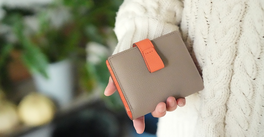 Atelier nuu 灰色皮革雙折短夾，橘色裝飾與內襯，由手持於米白色針織毛衣旁