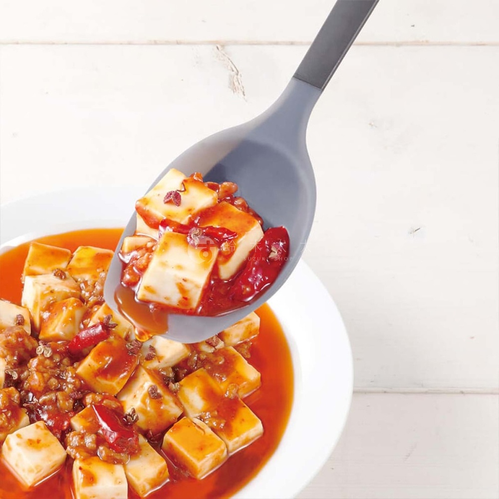 一個灰色矽膠料理匙，上面盛著麻婆豆腐，湯汁從湯匙邊緣滴落，背景是白色的木紋桌。