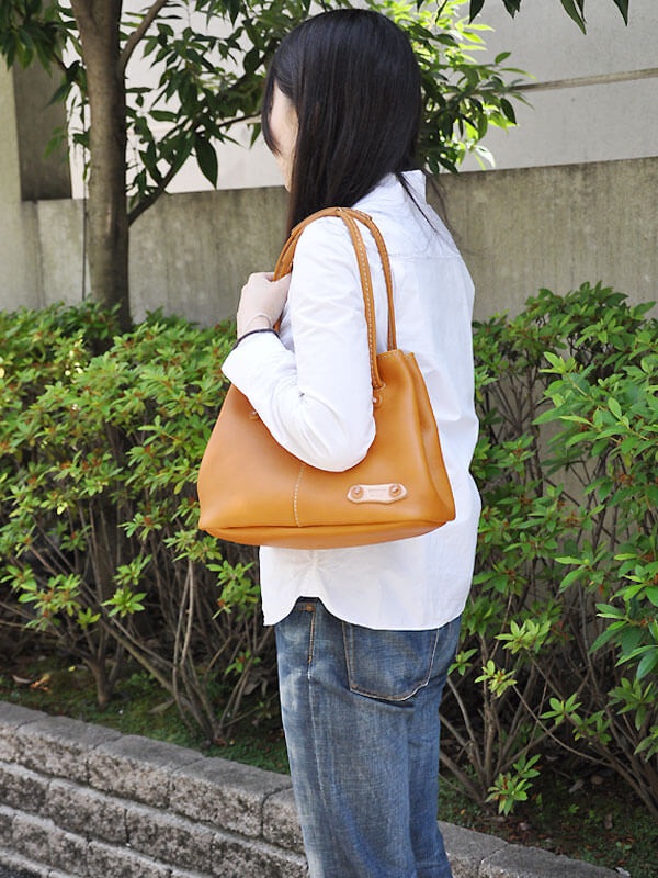 一位女性身穿白色襯衫和牛仔褲，肩上挎著一個棕褐色皮革托特包，包包上有兩個金屬扣環和縫線細節，背景為綠色植物和灰色牆壁。