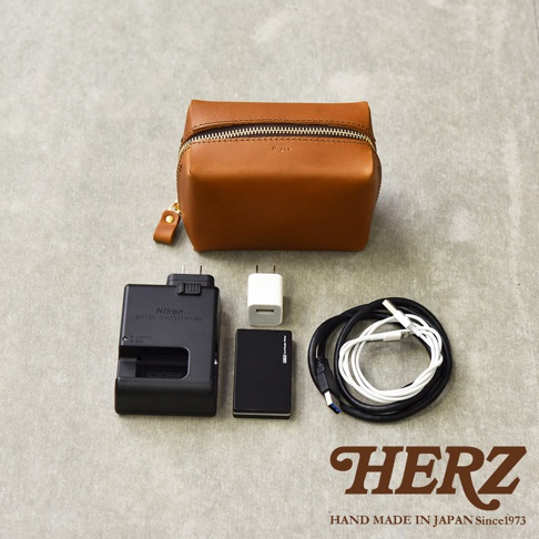 一個棕色皮革製的箱型收納包，旁邊擺放著相機電池充電器、一個黑色的小型裝置、一個白色充電頭和兩條充電線。