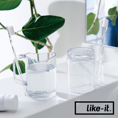 透明塑膠漱口杯，一個杯身有手柄，側邊有凹槽可放置牙刷，杯內裝有約半杯水。另一個漱口杯為圓柱形，側邊有凹槽可放置牙刷，杯內裝有約半杯水。兩杯皆置於白色檯面上，背景有綠葉植物。