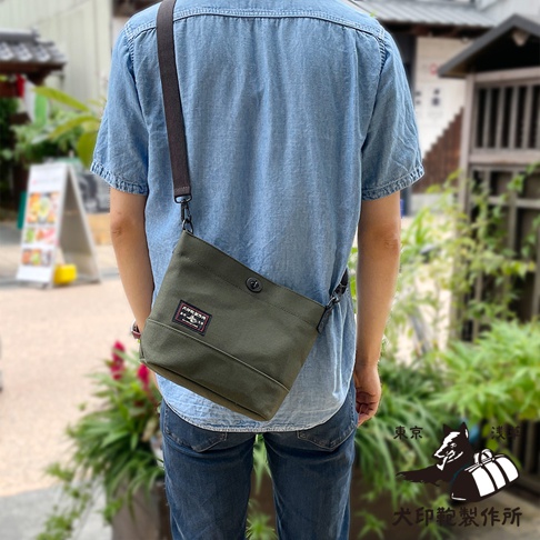 一位身穿藍色牛仔襯衫的男子，背對鏡頭，斜挎著一個綠色帆布側背包。背包上印有「東京 淺草 犬印鞄製作所」的標誌。