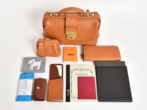 職人手作皮革兩用迷你口金單肩包 (CW-7)，展示於白色背景上。包包為棕褐色皮革材質，配有金色金屬配件和可調節揹帶。旁邊擺放著同系列的小零錢包、皮夾、卡夾、筆記本、護照套、濕紙巾和印有狗狗圖案的布巾。