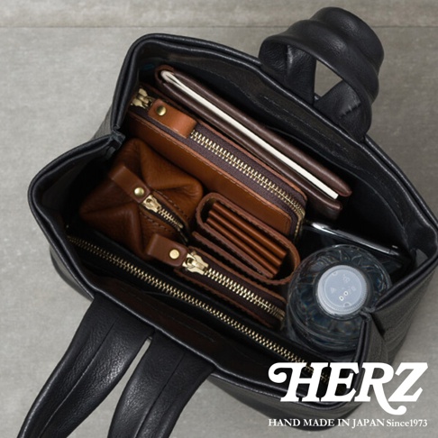 職人手作黑色皮革Organ單柄手提包，內部收納棕色皮革短夾、拉鍊零錢包與筆記本，並有寶特瓶。