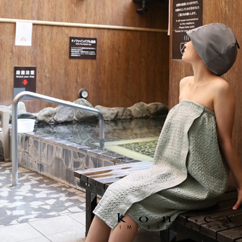 一位女士穿著淺綠色毛巾浴裙，頭戴灰色抗菌髮帶，坐在木凳上，身後是溫泉池。