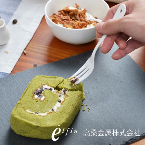 一雙日製白琺瑯水果甜點叉，叉柄帶有圓孔，一叉正叉著一塊綠色的抹茶瑞士捲蛋糕，蛋糕內餡有白色奶油和紅豆。