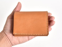 手持一個棕褐色皮革三折式迷你短夾，帶有明顯的縫線細節，簡潔的設計展現皮革的自然質感。