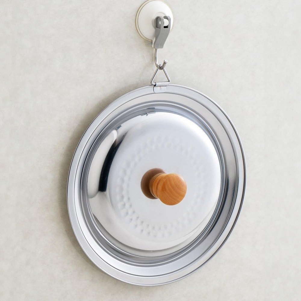 不鏽鋼鍋蓋，表面有蜂巢紋路，中央設有木質把手，透過掛鉤懸掛在牆壁上。