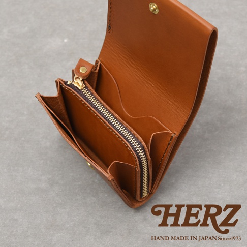 這款日本HERZ職人手作皮革短夾呈現溫潤的棕色質感，採用極簡俐落的雙折設計，內部配置拉鍊式零錢袋與實用的卡片收納隔層，右下角印有HERZ品牌商標與手作產地資訊。