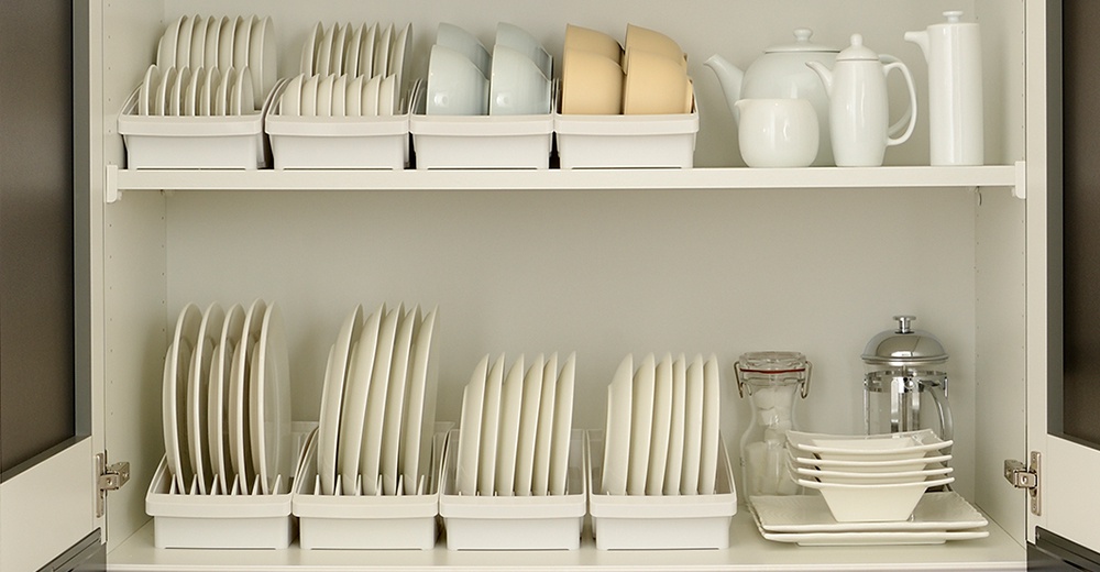 櫥櫃內擺放著整齊的餐具，上層有白色茶壺、奶盅、咖啡壺，以及淺藍色和淺黃色的碗，下層則有白色盤子和方形碗盤堆疊。