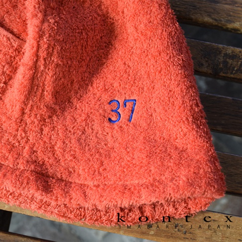 珊瑚橘色毛巾材質的桑拿帽，繡有藍色數字「37」，帽子邊緣有流蘇設計。
