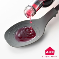 灰色量匙盛著深紅色液體，一瓶玻璃瓶傾斜，液體正從瓶口滴入量匙中。量匙內側有刻度標示。