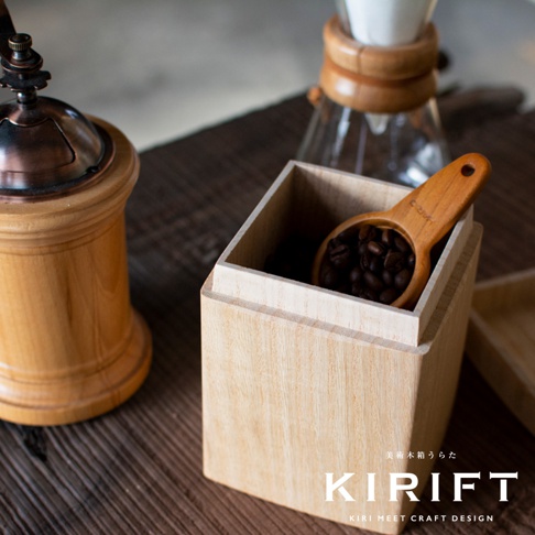 淺色桐木製咖啡豆保存盒，內有木製咖啡豆勺，盒內裝有深棕色咖啡豆。背景為咖啡磨豆機和濾滴咖啡壺。