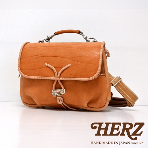 橘色皮革手作郵差包，有鹿角造型金屬扣環和抽繩閉合設計，搭配皮革提把和可調節斜背帶。