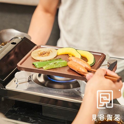 長方形的深棕色烤盤上烤著洋蔥、櫛瓜、青椒和兩根香腸，烤盤置於爐火上方，有人用筷子夾取香腸。