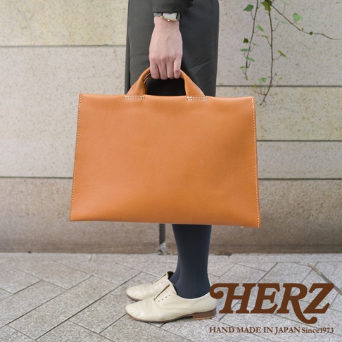 手提駝色簡約皮革公事包，搭配米白色牛津鞋，包包上有HERZ字樣。