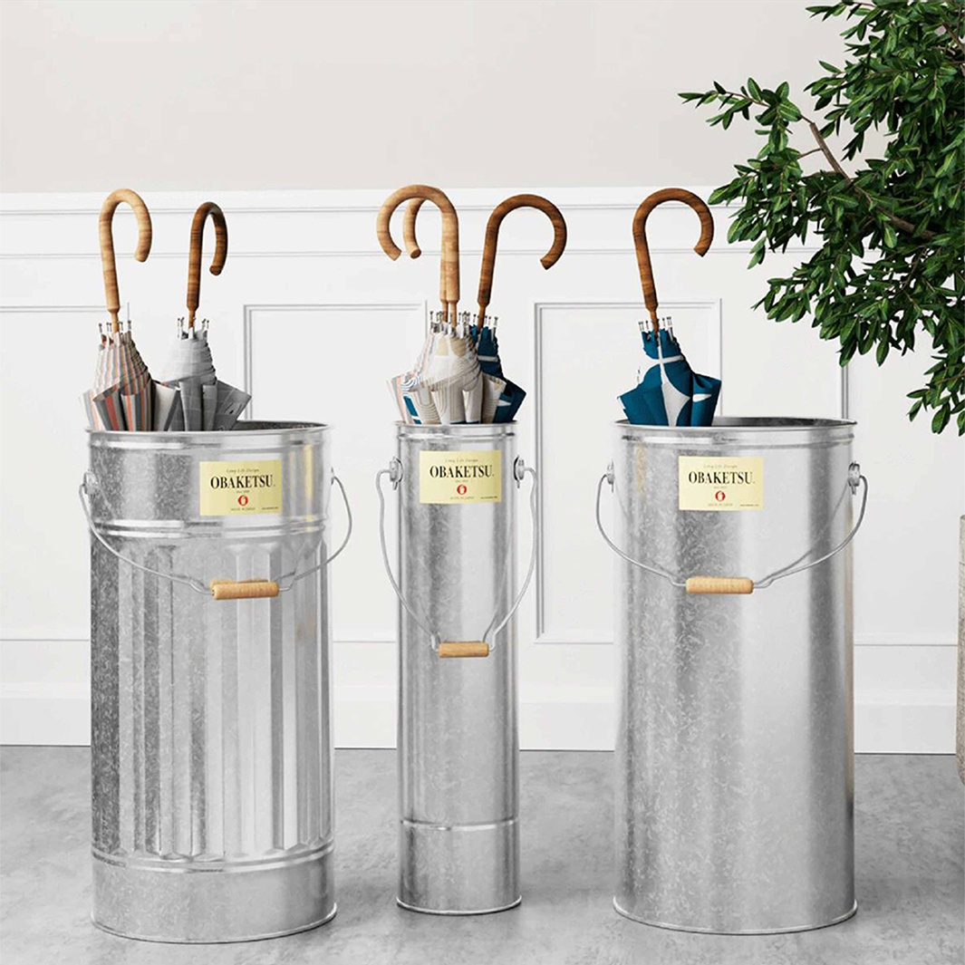 OBAKETSU 手工製傘桶，以銀色金屬製成，傘桶上印有 OBAKETSU 商標，搭配木質提把，展示傘架的簡約風格
