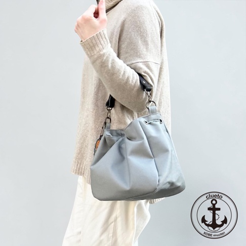 一位模特兒身穿米色針織上衣與白色寬褲，肩背著一只神戶clueto品牌的灰色防潑水BALON mini多用途手提包，包款呈現柔軟的束口造型並配有黑色皮革肩帶，右下角印有clueto KOBE since 2001的圓形錨圖案標誌。