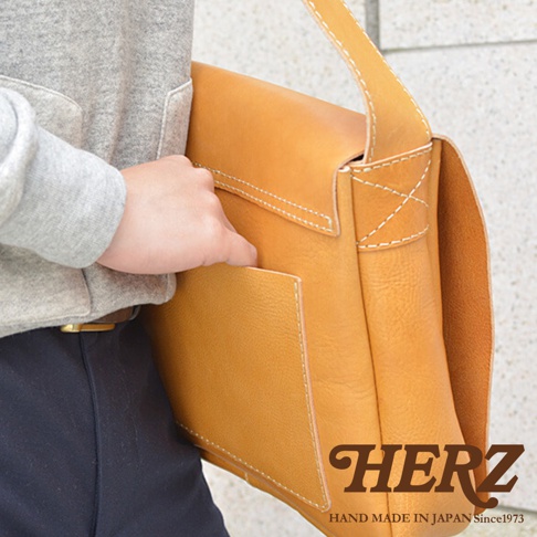 日本HERZ手工製作的棕褐色皮革斜背包，一個手伸入包包前方口袋。