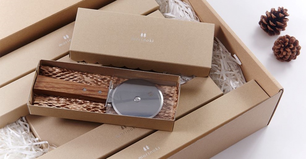 morinoki 比薩刀，銀色不鏽鋼刀輪，搭配木質握柄，裝於牛皮紙盒內，盒上印有 morinoki 商標，整體置於紙箱中，周圍填充白色碎紙絲，背景有兩個松果。