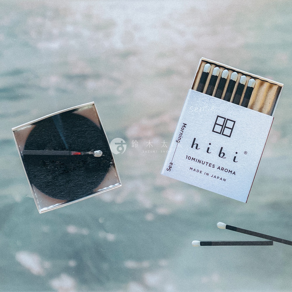一盒hibi香氛火柴開啟的狀態，展示了火柴排列在盒中的樣子以及一個放置在黑色圓形隔熱墊上燃燒的香氛火柴，另有兩根火柴平放在下方，背景呈現淡藍色的水波紋質感。