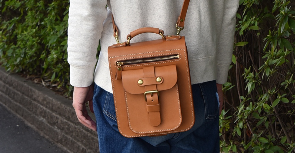 棕褐色皮革製成的立體口袋兩用單肩包，配有拉鍊主隔層、翻蓋前袋、金屬扣和可調節肩帶，由手提把連接。