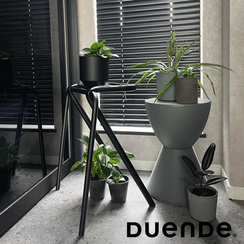 黑色金屬框架的吧檯椅，椅面上有盆栽植物，旁邊有一個灰色階梯狀的置物架，上面也有盆栽植物，背景是百葉窗和落地鏡。