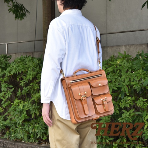 手工皮革三用後背包，採用棕色皮革材質，設有頂部提把、可拆卸肩帶、拉鍊主袋及多個前置口袋，口袋蓋以金屬扣環固定。